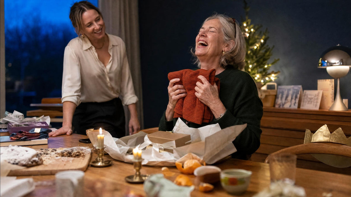 Oma lacht met dichte ogen terwijl ze een roestkleurige wafelhanddoek uit een kerstpakketje tegen zich aan houdt, haar dochter kijkt toe aan de kersttafel bij kaarslicht — wasparfum als kerstcadeau dat al ruikt bij het openen