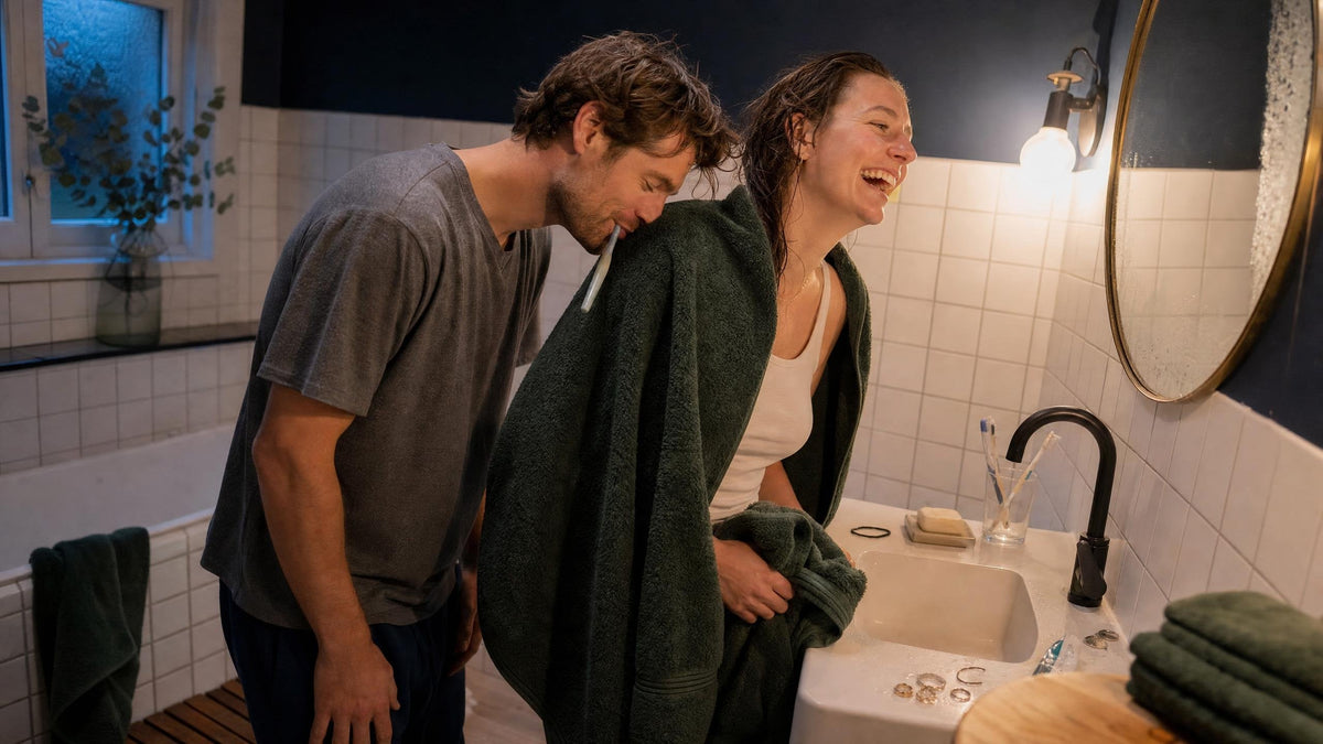 Stel in de badkamer op zaterdagavond: zij lacht met een vers gewassen groene badhanddoek om haar schouders terwijl hij met dichte ogen aan de handdoek ruikt — handdoeken wassen en lekker laten ruiken