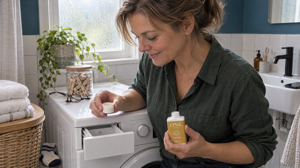 Vrouw houdt een dopje wasparfum boven de wasmiddellade van haar wasmachine, met het open flesje First Light in haar andere hand